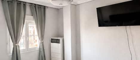 A bedroom with a bed, curtains, a ceiling fan, an air conditioning unit, and a mounted TV.