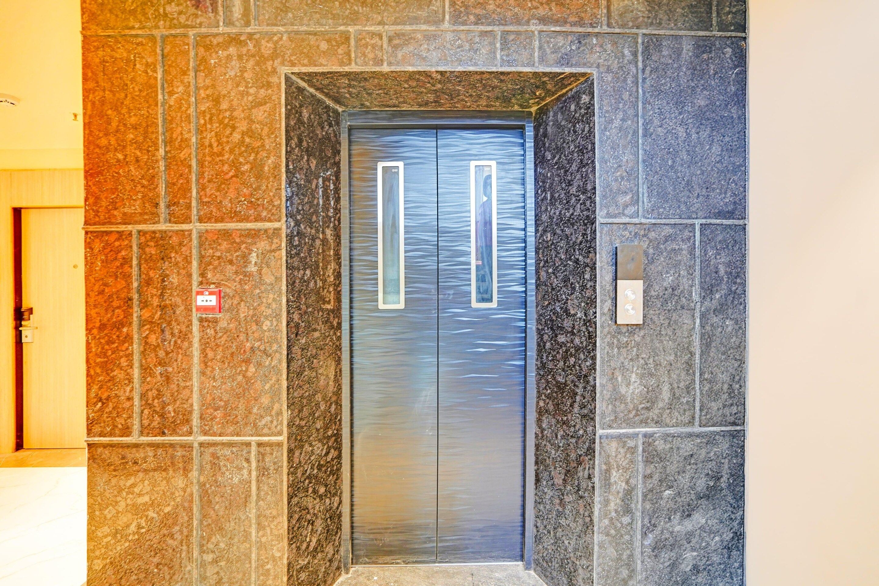 An elevator with a metallic door and a rectangular handle, set in a stone wall with a door on the left.