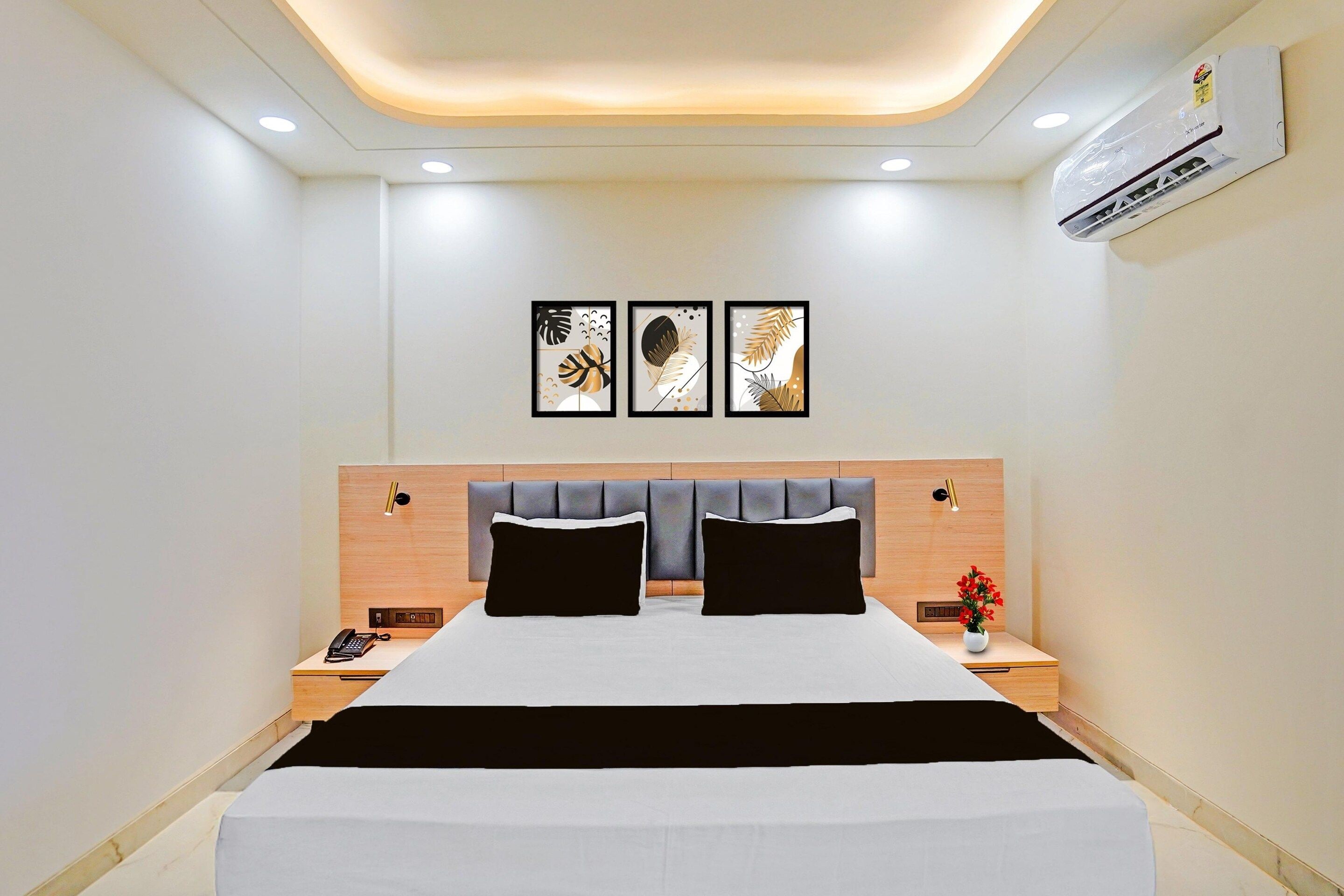 A modern hotel room with a bed, bedside tables, a nightstand, a wall-mounted air conditioner, and three framed artworks above the bed.