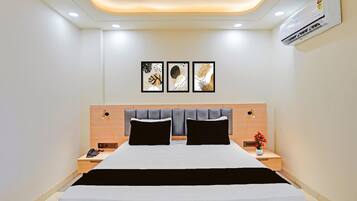 A modern hotel room with a bed, bedside tables, a nightstand, a wall-mounted air conditioner, and three framed artworks above the bed.
