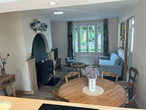 A dining area with a round wooden table, chairs, and a vase with flowers. There is a fireplace with a mantel, a sofa, and a television in the background. - La maison bleue (Saint-Valery-sur-Somme)