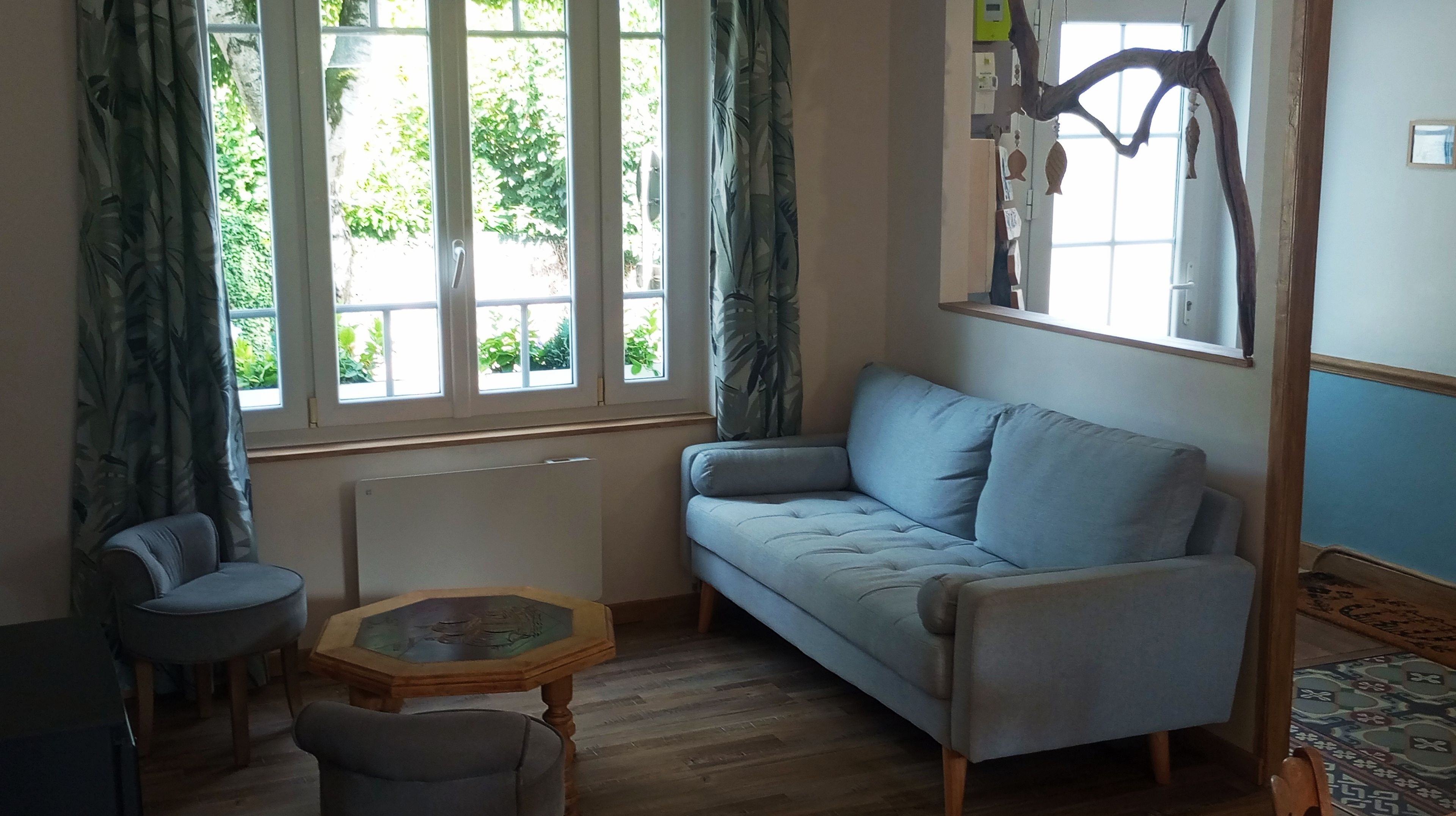 Une pièce comprenant un canapé, une petite table et une chaise. On y trouve des fenêtres avec des rideaux, un radiateur et une étagère fixée au mur, ornée d’objets décoratifs.