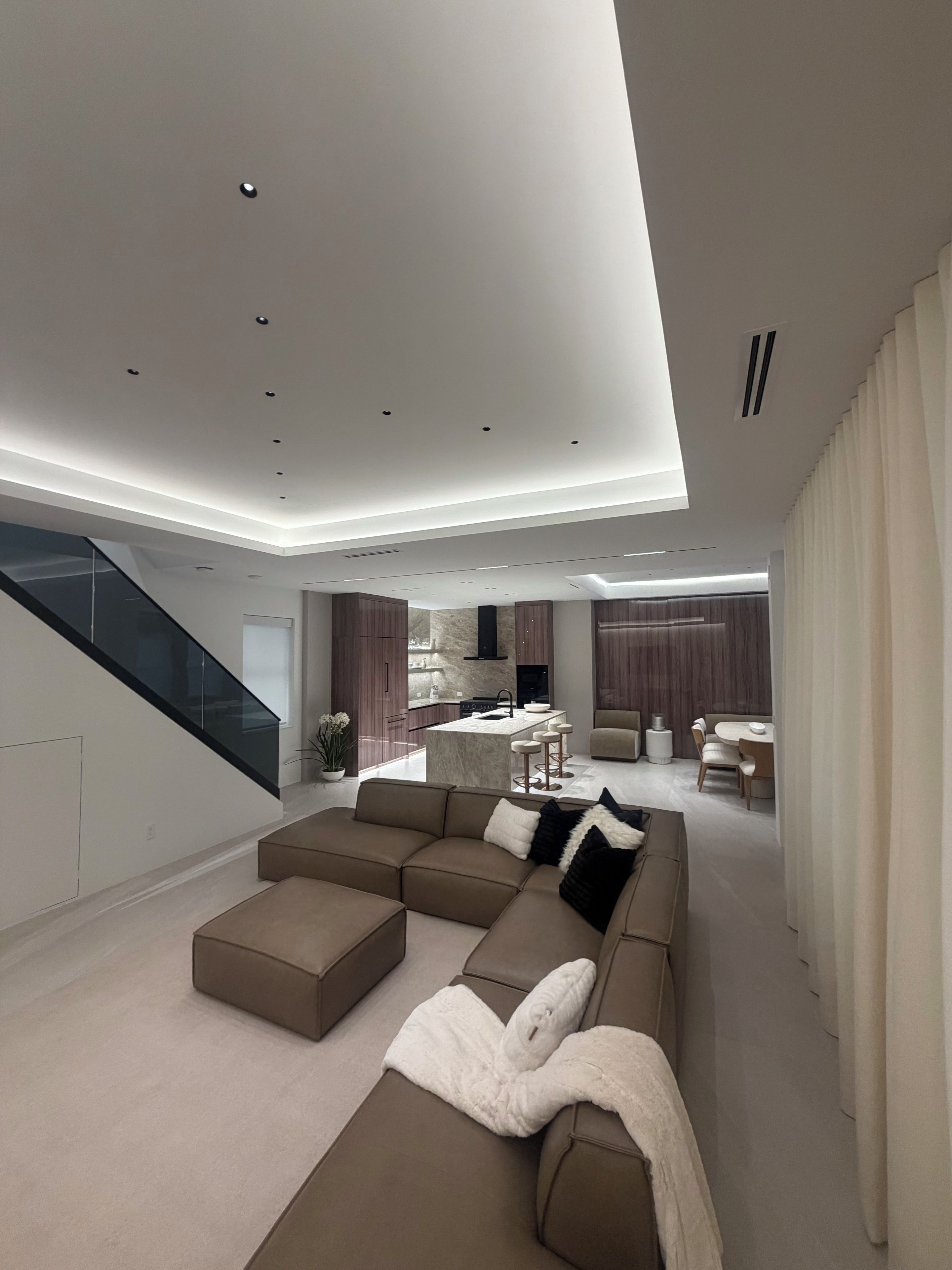 A modern living room with a sectional sofa, a kitchen area, and a staircase.