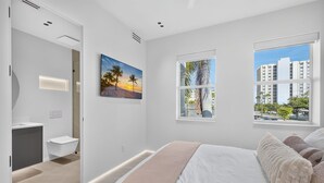 Una camera da letto moderna con un letto grande, un bagno con vasca e vista su palme e edifici.