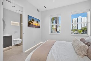 Une chambre moderne avec un grand lit, une salle de bain avec baignoire et une vue sur les palmiers et les immeubles.