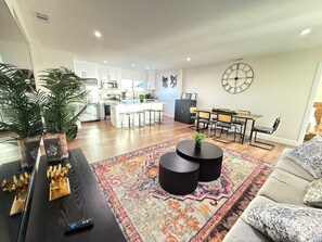 A modern living room with a sofa, a coffee table, and a rug. There is a kitchen in the background with white cabinets and a dining area with a table and chairs.