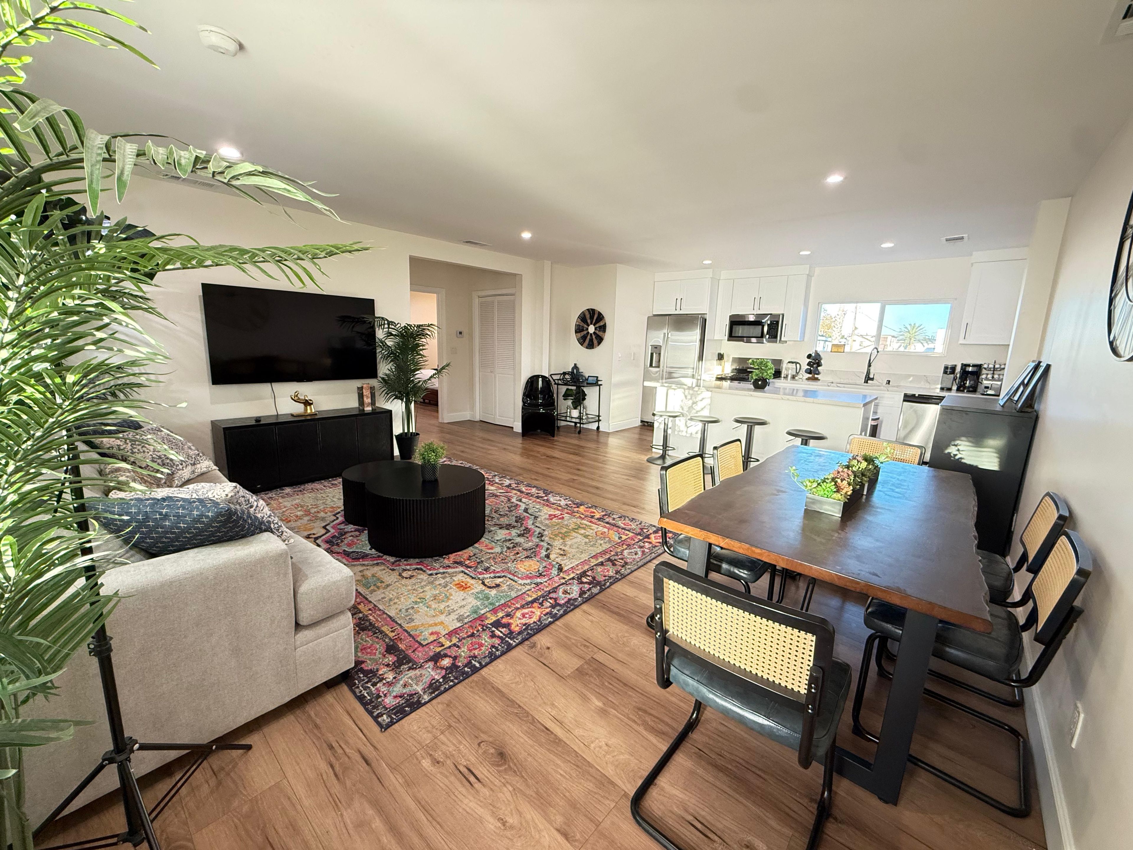 Ein moderner Wohnbereich mit Sofa, Couchtisch, Esstisch und Stühlen. Ein Flachbildfernseher ist an der Wand montiert. Der Raum verfügt über Holzdielen und einen farbenfrohen Teppich.