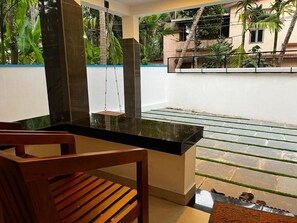 Un espacio de estar moderno al aire libre con una mesa de vidrio y sillas de madera, una hamaca colgante y vistas a una zona residencial con palmeras.