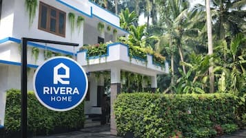 A hotel entrance with a sign reading 'RIVERA HOME,' surrounded by lush greenery and palm trees.