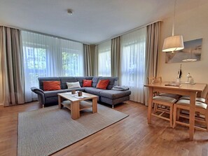 A modern living room with a grey sectional sofa, a wooden coffee table, a dining table with chairs, and sheer curtains on the windows. - Holiday Apartment 3.02 Island Beach (Heringsdorf)