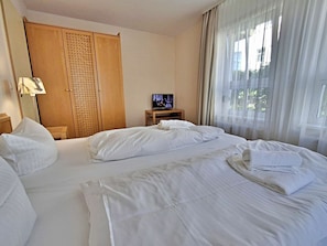 Ein Hotelzimmer mit zwei Betten, einem Fernseher auf einem Holzboden, einem Kleiderschrank und einem Fenster mit durchsichtigen Vorhängen.