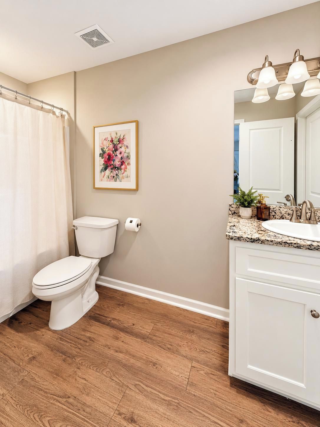 A bathroom with a toilet, a shower curtain, a framed floral artwork, a sink with a mirror, and a vanity cabinet.