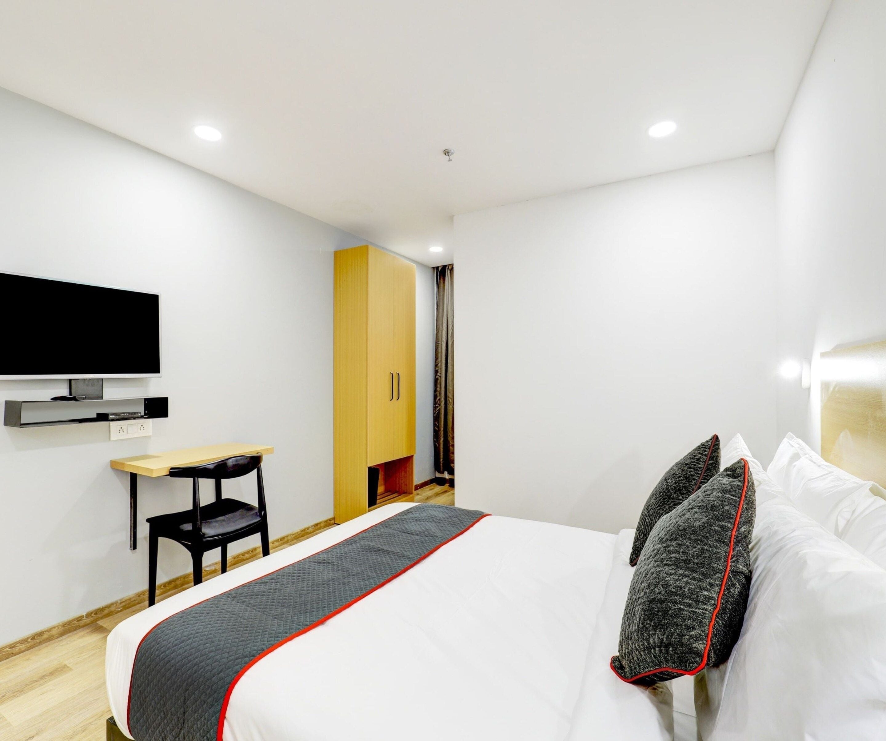 A modern hotel room with a bed, a desk, a chair, a television, and a wardrobe.