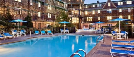 A pool area with lounge chairs and umbrellas, surrounded by a building with brick walls and multiple windows.