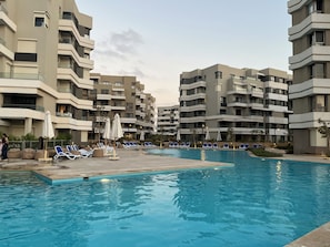 Una piscina con sillones de descanso y sombrillas, rodeada de edificios residenciales de varios pisos.