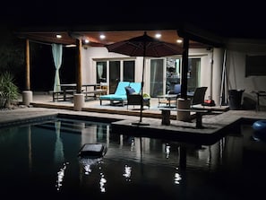 A poolside area with lounge chairs, a table, and a covered seating area at night.