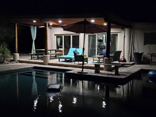 A poolside area with lounge chairs, a table, and a covered seating area at night.