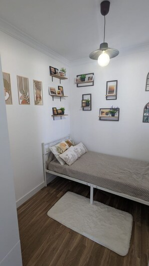 Une petite chambre à coucher, bien rangée, avec un lit simple, un tapis blanc et des étagères murales ornées de photos encadrées et de plantes.