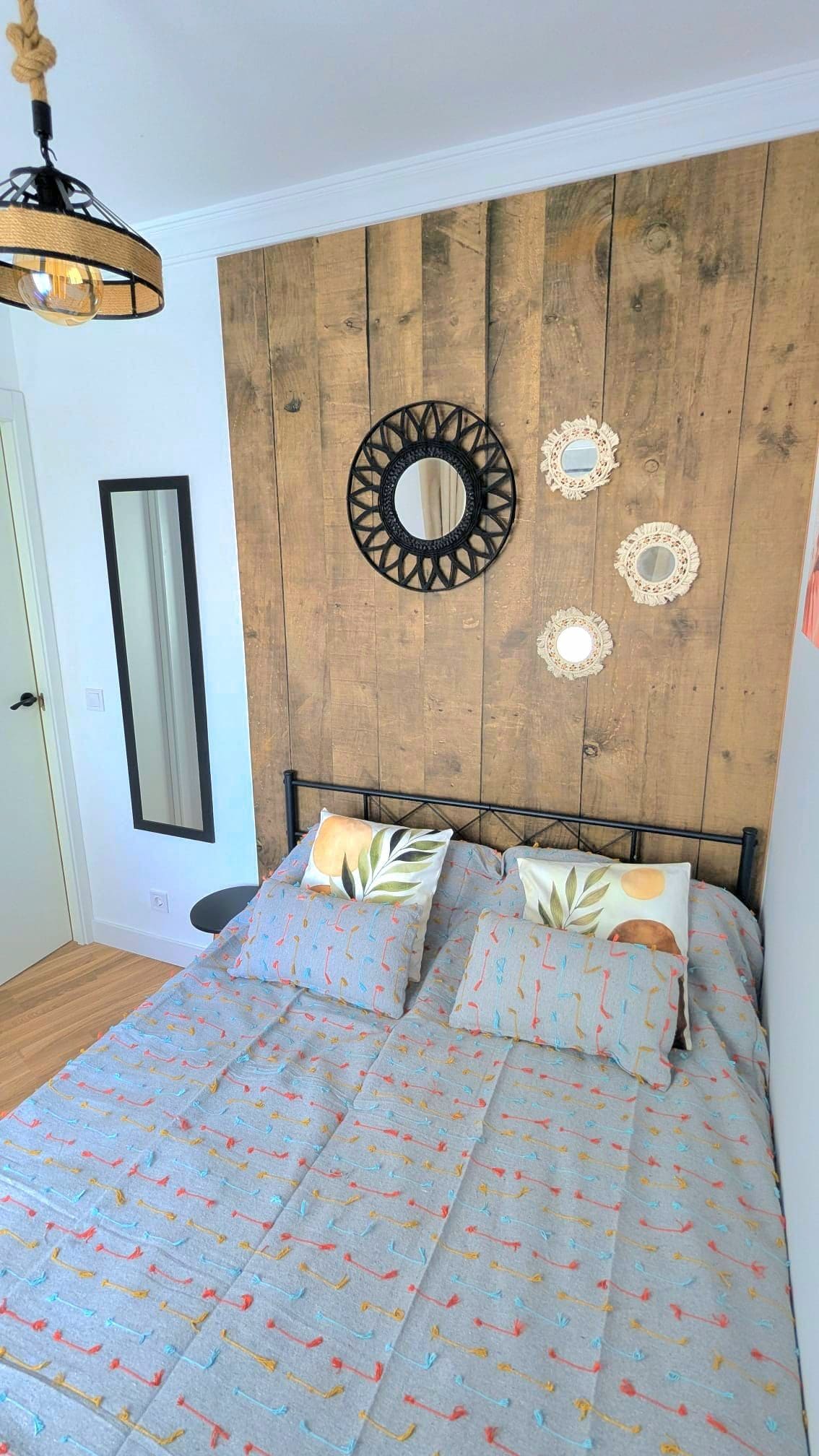Quarto com cabeceira de madeira, cama com roupa de cama estampada, luminária pendente e espelhos decorativos na parede.