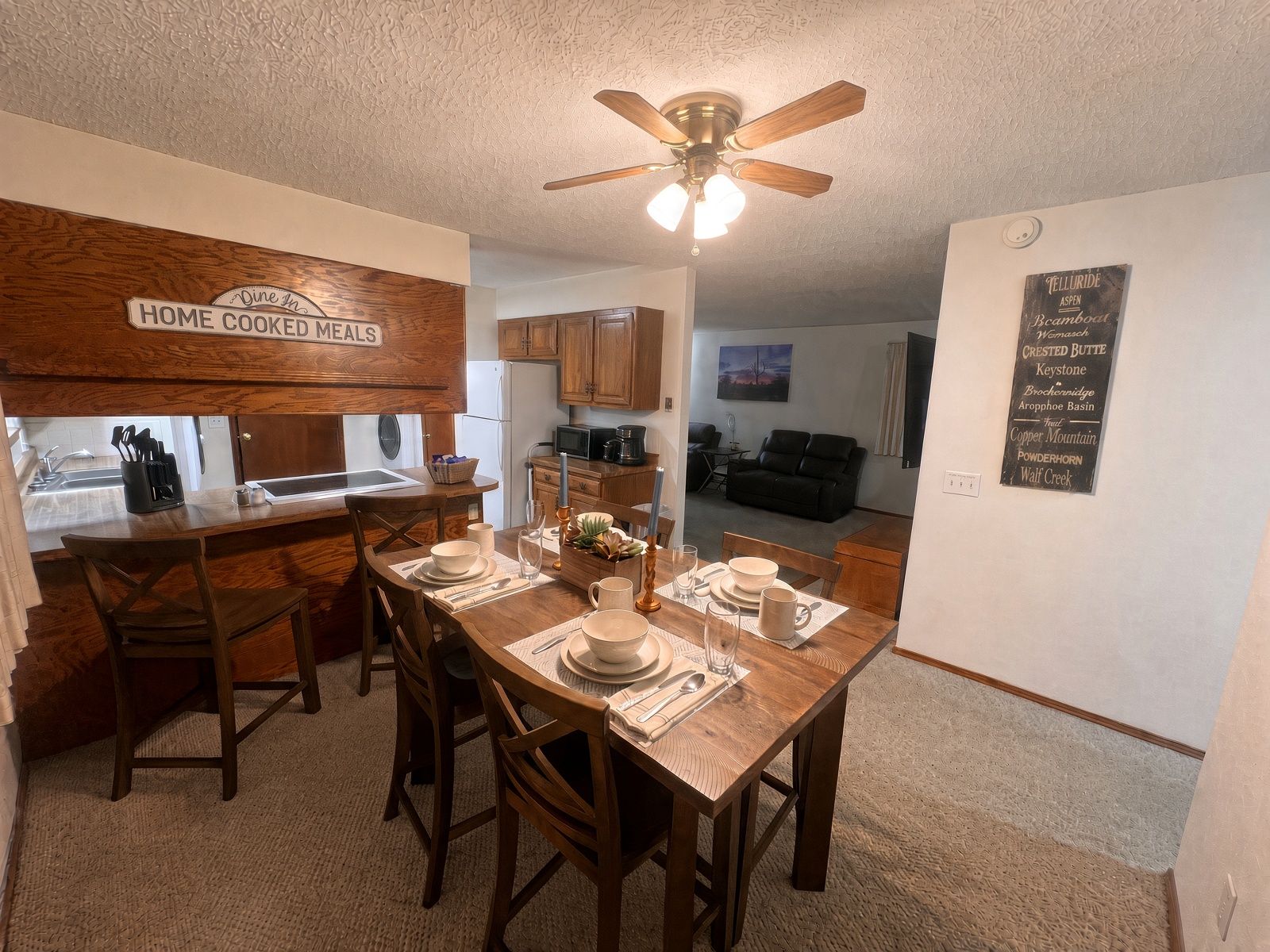 A dining area with a wooden table set for four, a ceiling fan, and a kitchen in the background.
