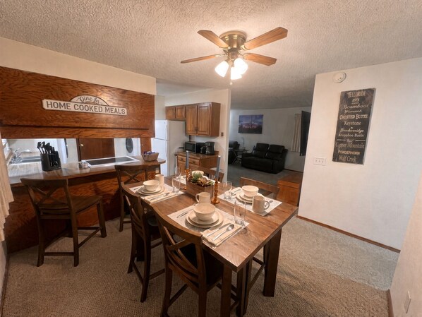 A dining area with a wooden table set for four, a ceiling fan, and a kitchen in the background.