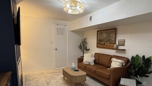 Un salon avec un canapé marron, une table basse en bois, un tapis à motifs, une plante en pot et un tableau accroché au mur.
