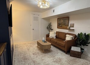Ein Wohnzimmer mit braunem Sofa, einem Holztisch, einem gemusterten Teppich, einer Topfpflanze und einem Gemälde an der Wand.