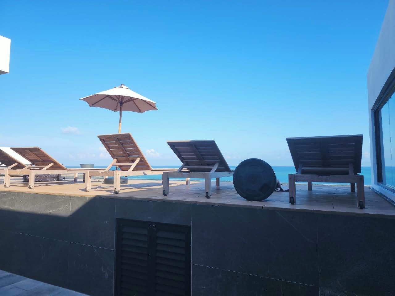 Eine Dachterrasse mit Liegestühlen und einem großen Sonnenschirm, mit Blick auf einen klaren, blauen Himmel und eine entfernte Küstenlinie.