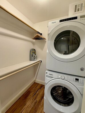 Une buanderie équipée d’un lave-linge et d’un sèche-linge superposés, d’étagères en bois et d’un balai.