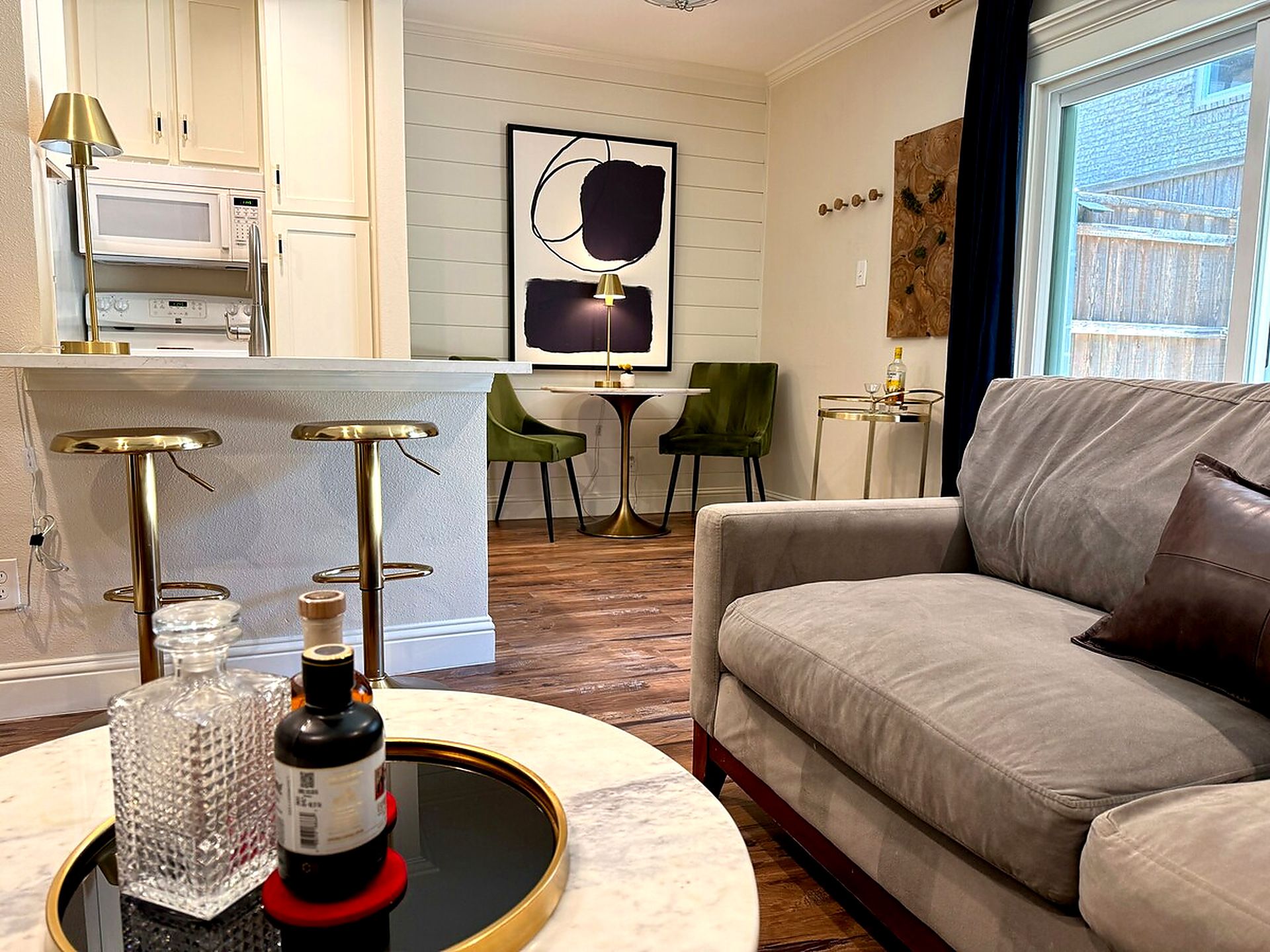 Un salon moderne avec un canapé gris, une table basse sur laquelle se trouvent une bouteille et un verre, et un coin cuisine avec des meubles blancs et un four à micro-ondes.