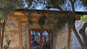 Una pérgola de madera con una mesa y sillas, rodeada de arena y plantas.