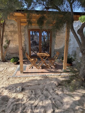 Eine hölzerne Pergola mit Tisch und Stühlen, umgeben von Sand und Pflanzen.