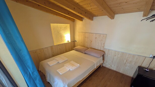Una cama individual con sábanas y toallas blancas, un cabecero de madera, una mesita de noche con lámpara y una ventana con cortinas azules.