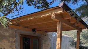Una pérgola de madera con una mesa y sillas, una planta en maceta y un camino de piedra que lleva a una puerta de cristal.