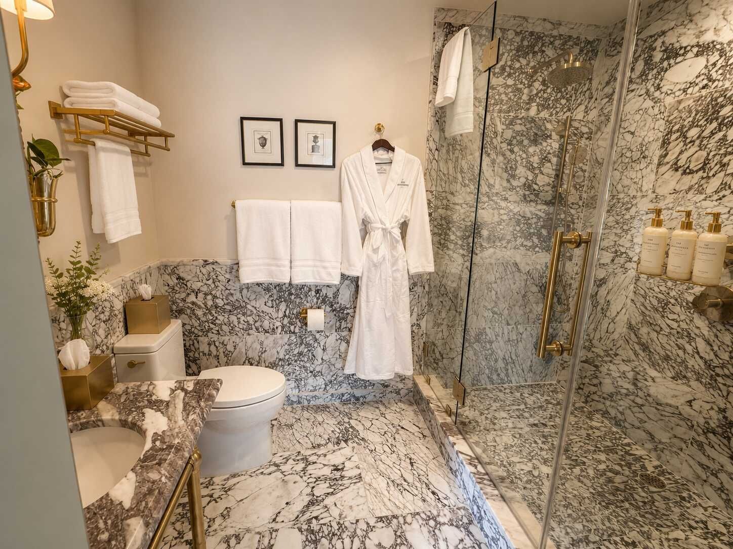 A marble bathroom with a toilet, sink, and shower. There are towels and a robe hanging on hooks.