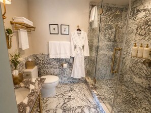 Een marmeren badkamer met toilet, wastafel en douche. Handdoeken en een badjas hangen aan haken.