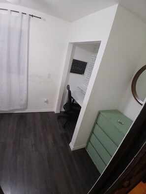 A room with a dark wooden floor, a green cabinet, a black office chair, a white curtain, and a small window with a black frame.