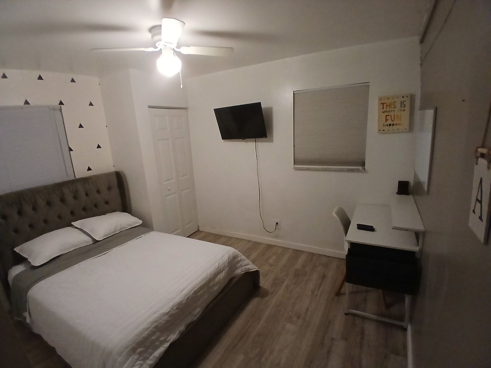 Un dormitorio con cama, escritorio, televisor, ventilador de techo y ventana.