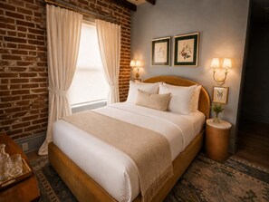 Ein Hotelzimmer mit Backsteinwand, einem großen Bett mit weißer Bettwäsche, zwei Wandlampen und einem Fenster mit Vorhängen.