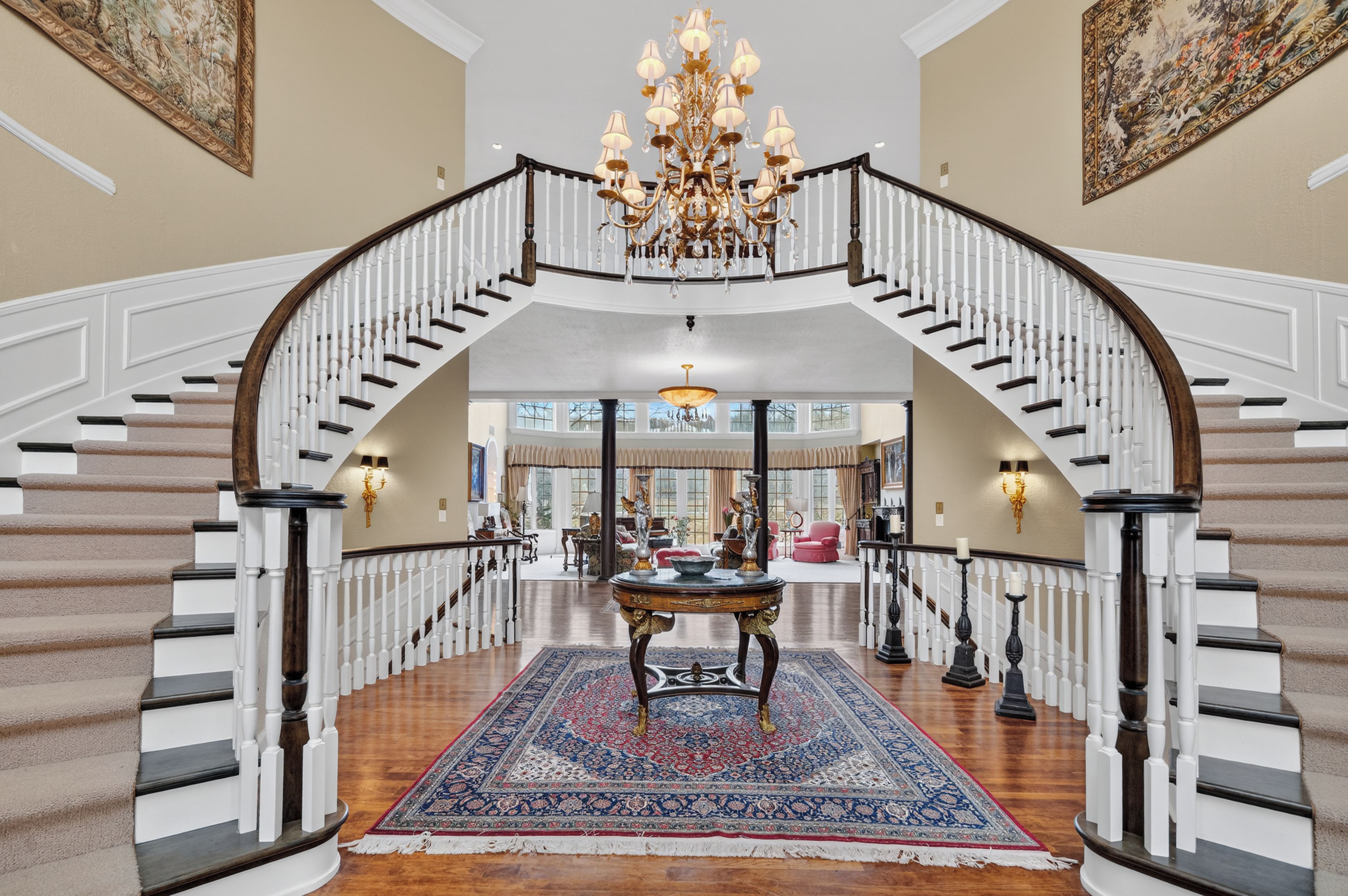 A grand staircase with a chandelier, a large rug, and a view into the living area.