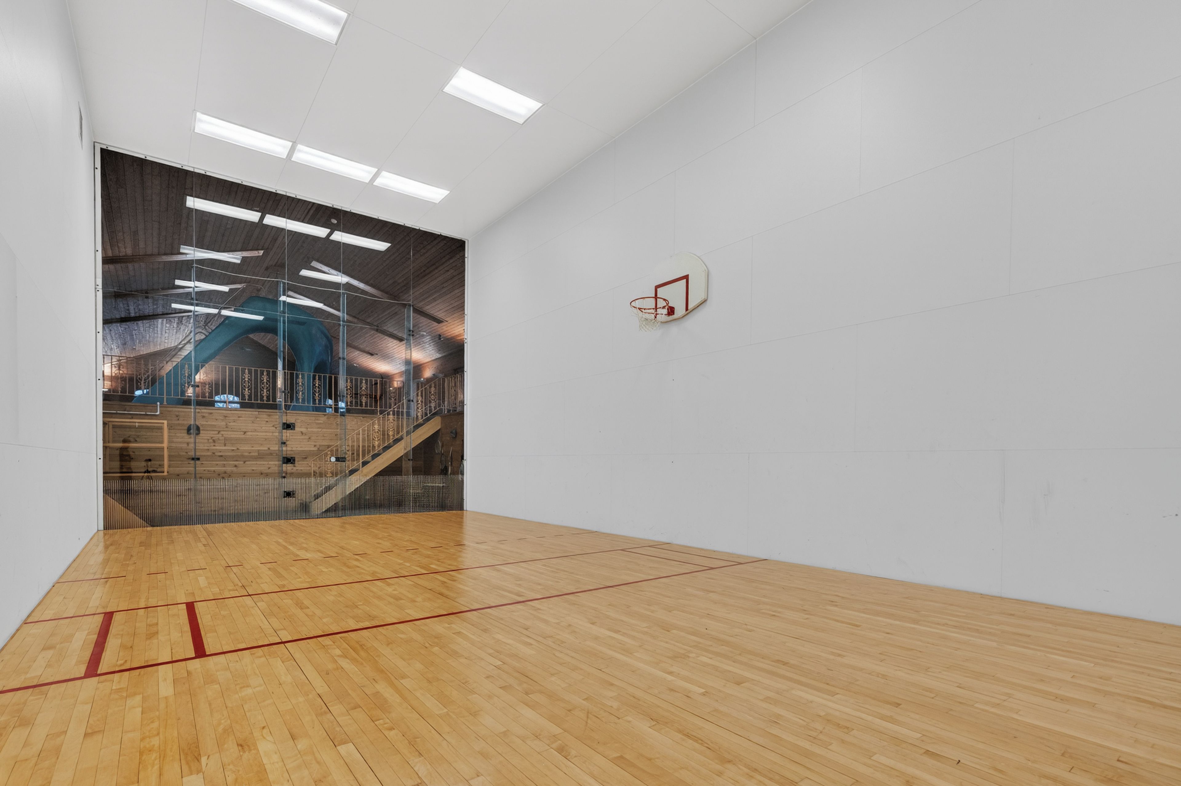 A spacious indoor gym with a wooden floor, a basketball hoop, and a large window overlooking a wooden staircase.