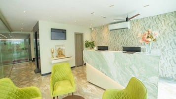 A reception area with a marble countertop, a flat-screen TV, and a decorative plant. There are two green upholstered chairs and a small round table.