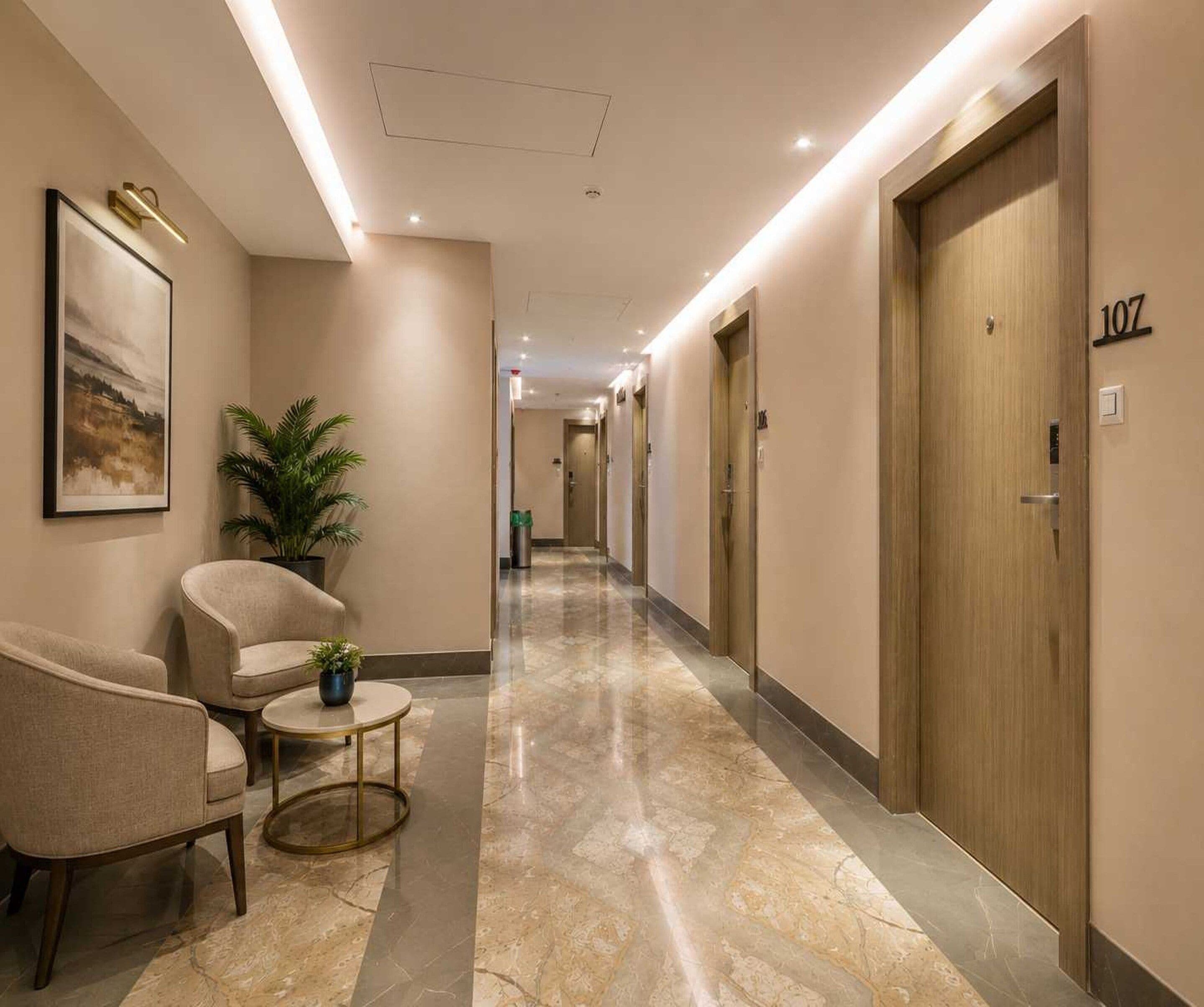 A hotel hallway with a marble floor, a seating area with chairs and a small table, and doors on the right side.