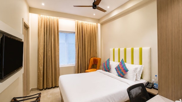 Quarto de hotel com cama, escrivaninha, cadeira, televisão, ventoinha no teto e cortinas.
