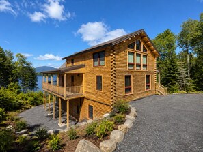 A log cabin with a balcony, a gravel driveway, and a forested backdrop.