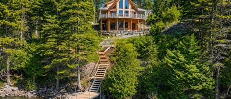 A cabin with a wooden deck leading to a dock over water, surrounded by dense forest.