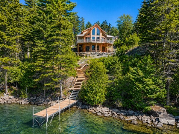 A cabin with a wooden deck leading to a dock over water, surrounded by dense forest.