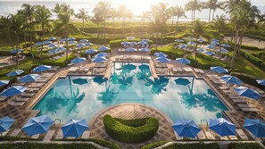 Un espace piscine dans un complexe hôtelier, doté de nombreuses chaises longues et de parasols, entouré de palmiers et offrant une vue sur l’océan.