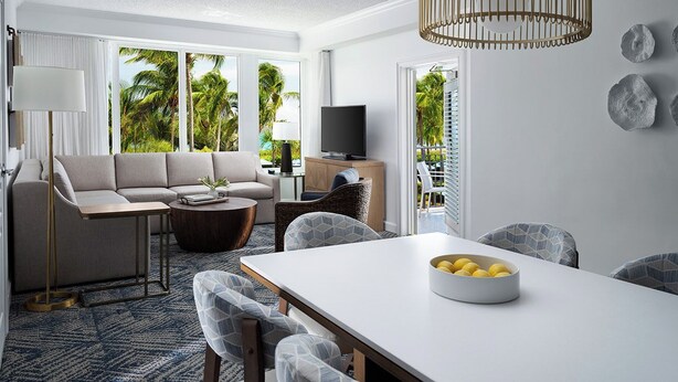 Una sala moderna con sofá, mesa de centro y mesa de comedor. Hay un televisor y una ventana con vistas a palmeras.