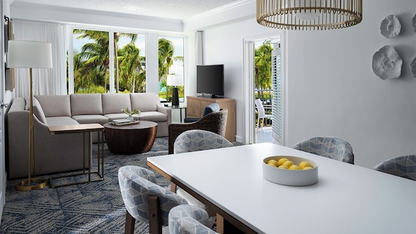 Un salon moderne avec un canapé, une table basse et une table à manger. On y trouve également un téléviseur et une fenêtre donnant sur des palmiers.